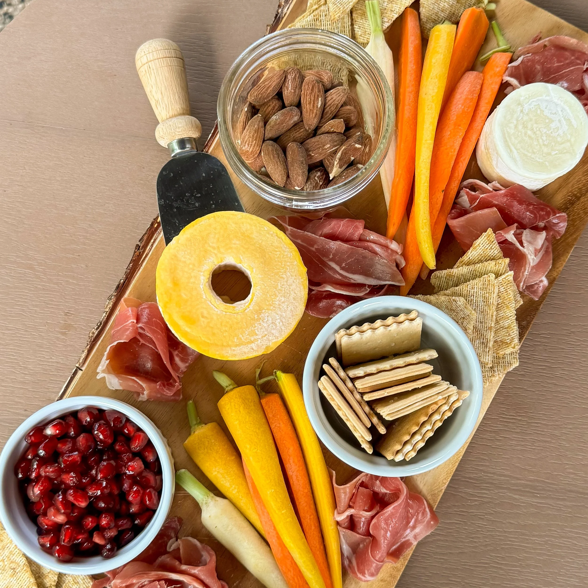 A wooden board with various food items including almonds in a jar, cherry tomatoes in a bowl, assorted cheeses, carrot sticks, cherry ham slices, and a small bowl of crackers.