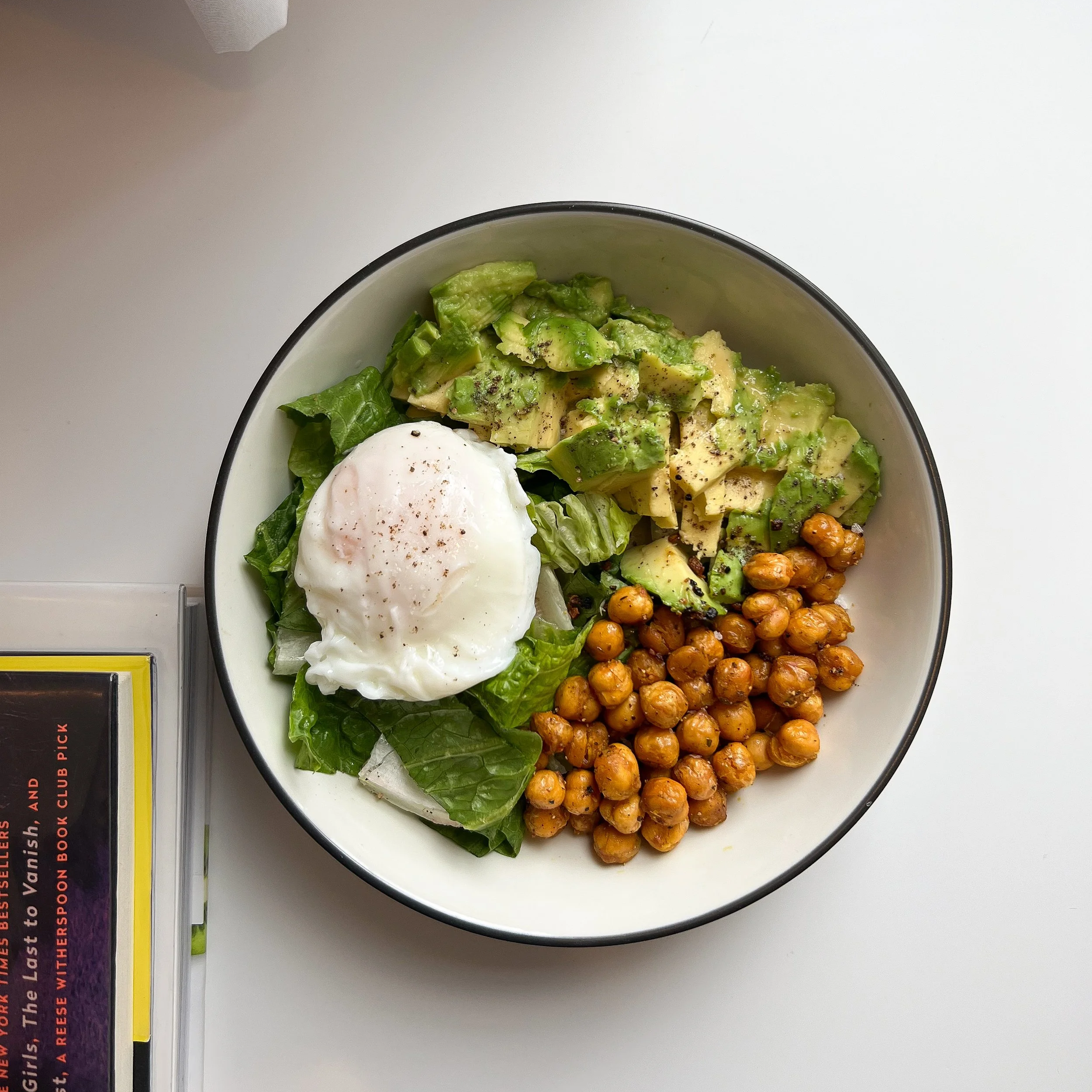 A bowl with chopped avocado, a poached egg, roasted chickpeas, and green leaf lettuce on a white surface.