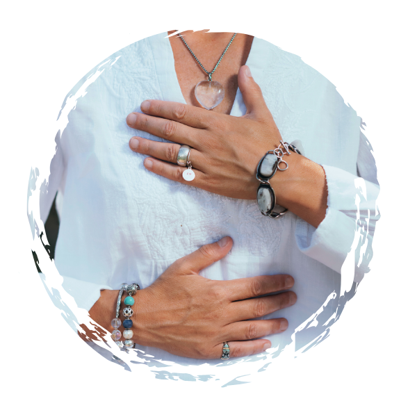 A woman wearing a white shirt with multiple jewelry pieces, including a heart-shaped pendant necklace, several rings, and bracelets with various beads and charms, with one hand placed on their chest and the other on their stomach receiving Reiki.