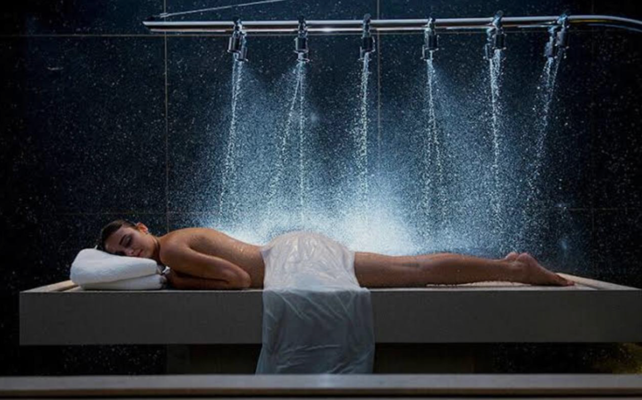 A woman lying on a bed in a spa, receiving a water massage from a showerhead overhead in a dark tiled room.