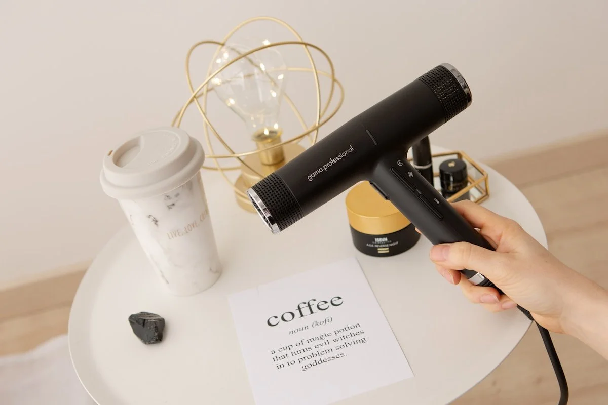 Person using a black handheld hair dryer on a white table. On the table are a disposable coffee cup, a black rock, a small candle in a container, a decorative lamp with a light bulb and gold wire frame, and a printed paper about coffee.