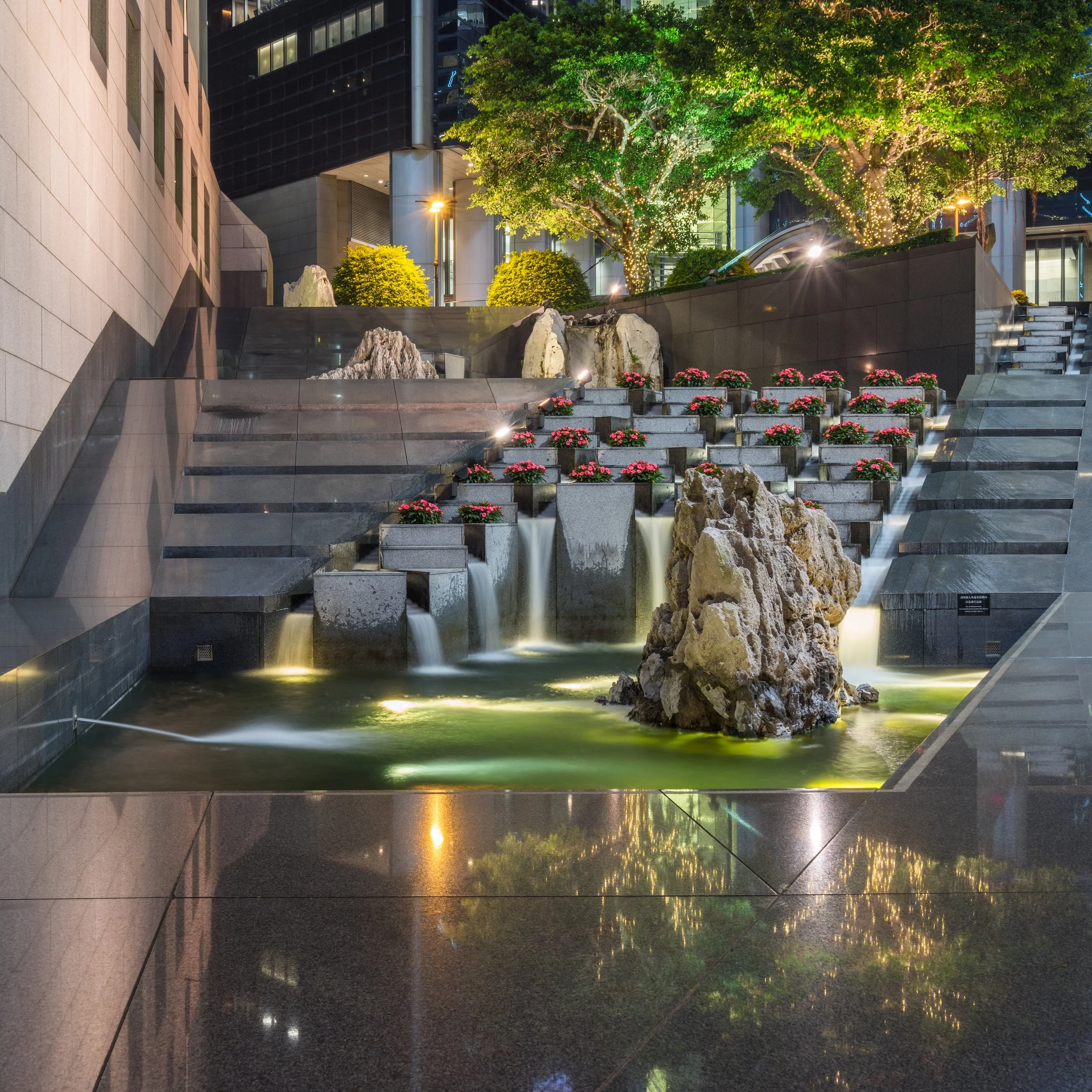 Nighttime scene of a modern cityscape with illuminated trees, water feature, and reflective ground surface.