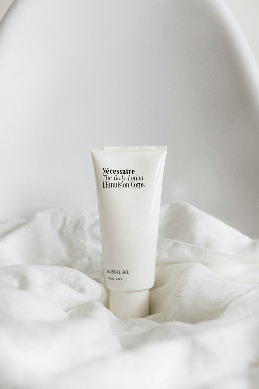 A white tube of Nécessaire The Body Lotion on a white fabric surface with a plain white background.