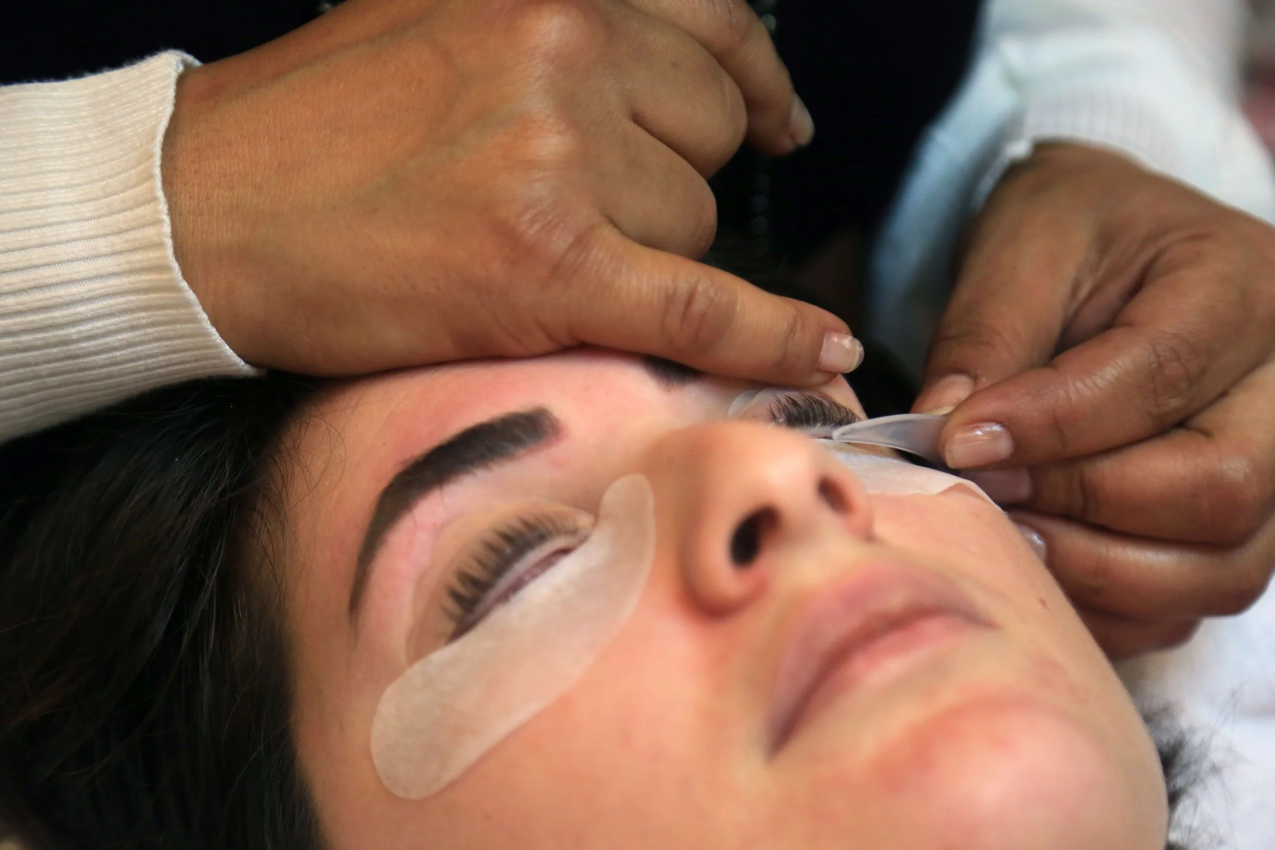 Close-up of a person receiving an eyelash extension procedure, with technician's hands carefully applying eyelash extensions to the client's natural lashes.