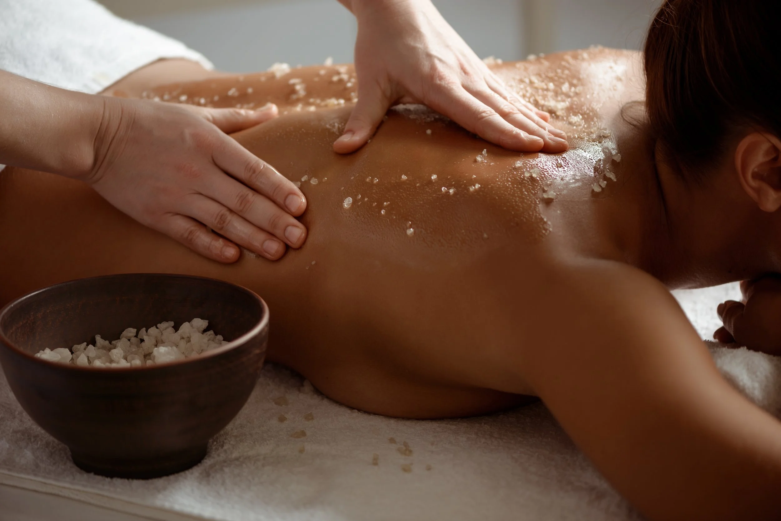 A person receiving a back massage with sea salt sprinkled on their back, while another person applies massage pressure. A bowl of sea salt and a bowl of oil are nearby.