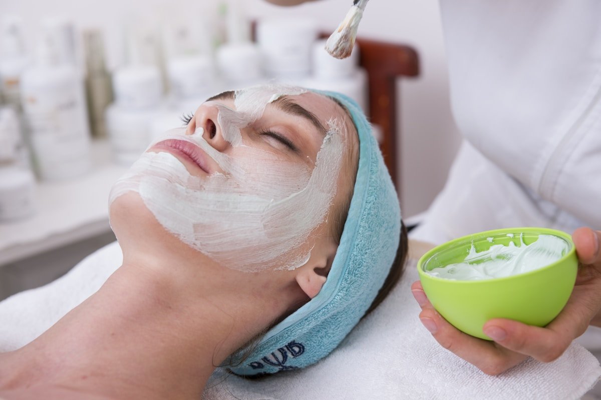 A woman receives a facial treatment at a spa, with a skincare mask being applied to her face by a professional.