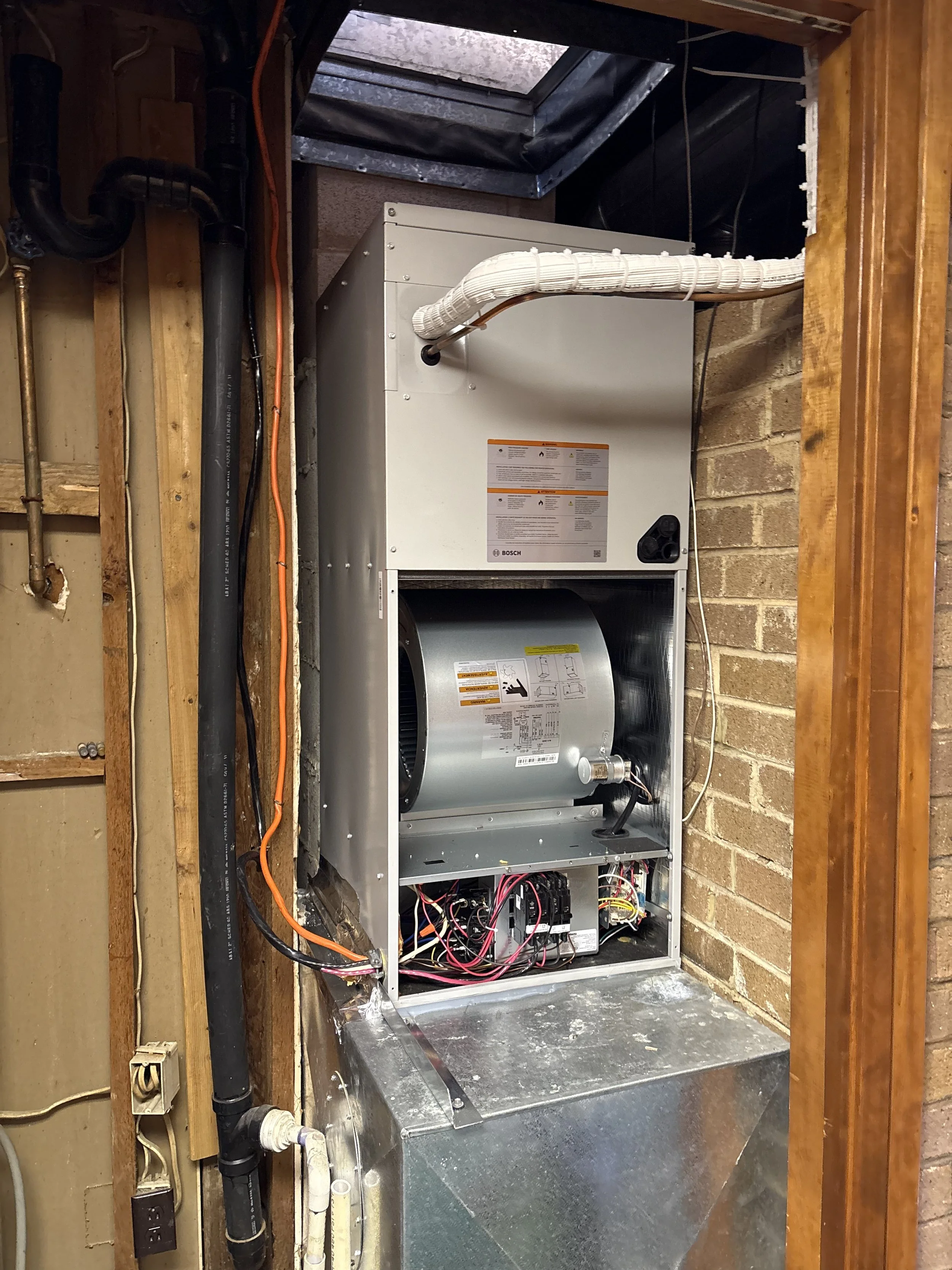 Interior view of an HVAC furnace unit with wiring and ductwork, located in a basement with brick walls and wooden framing.