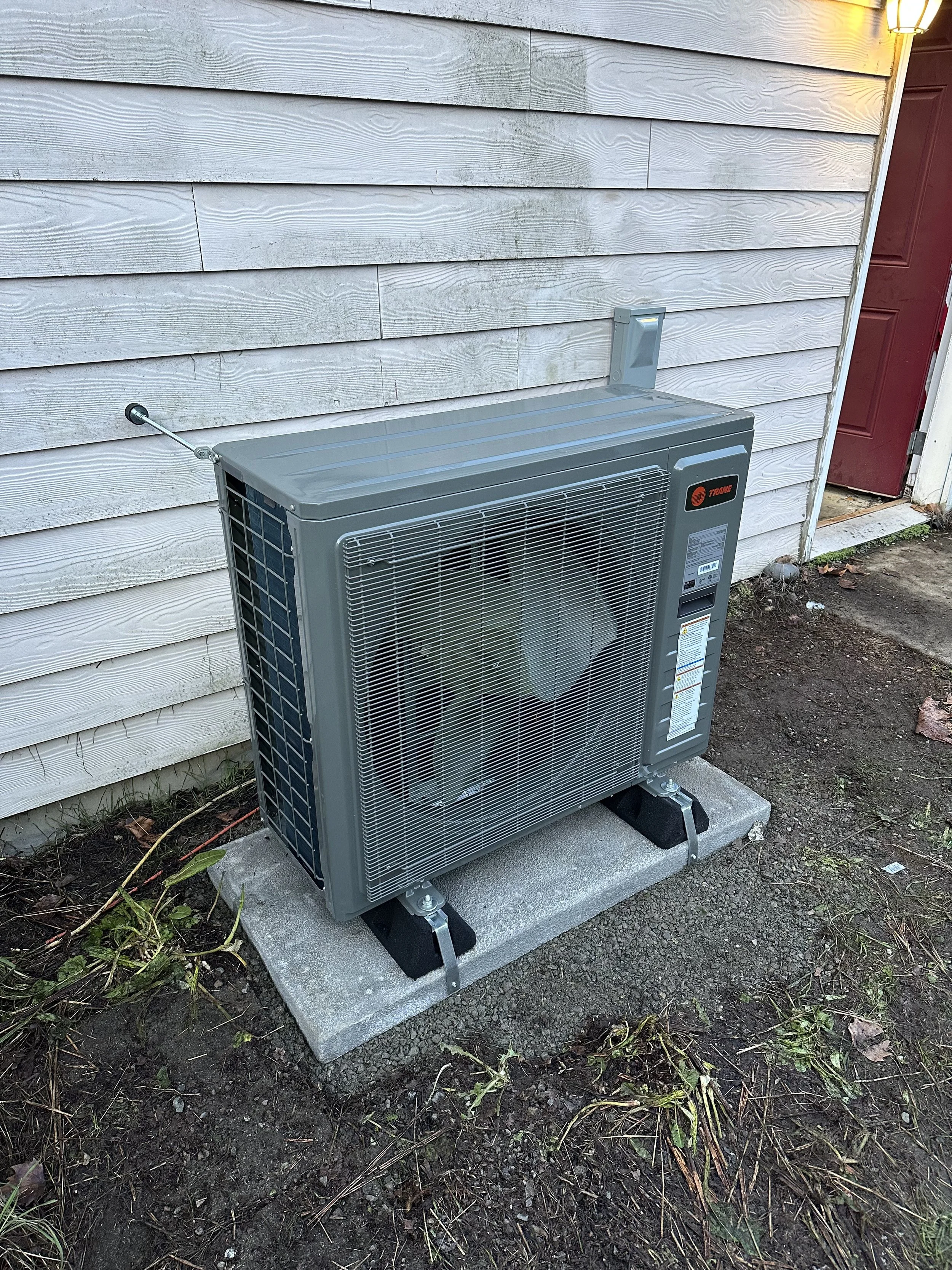 An outdoor air conditioning unit installed on a concrete pad next to a house with weathered horizontal siding. The unit has a metal grille and is connected to the house with electrical wiring. There is soil and some plants around the unit.