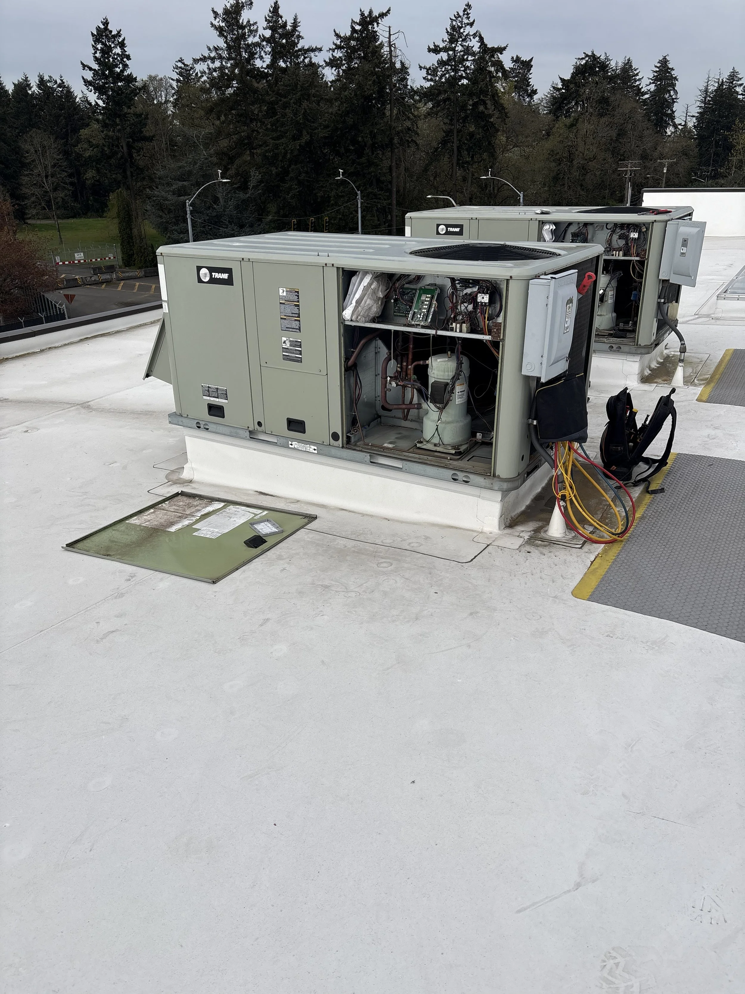 Two rooftop HVAC units with open panels revealing internal components, situated on a flat white rooftop with surrounding trees and utility poles in the background.