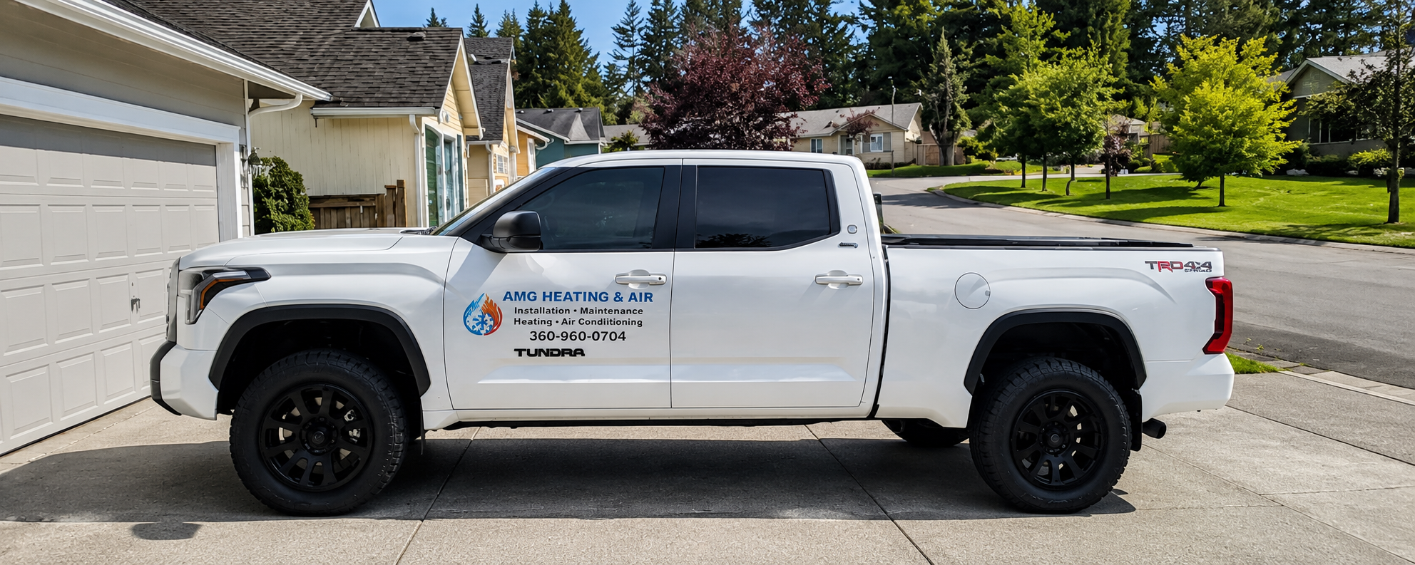 White pickup truck parked in a driveway in a suburban neighborhood. The truck has logo and contact info for AMG Heating & Air on the side, including a snowflake, flame, and water drop icon, with phone number 360-960-0704. The background features houses, trees, and a clear sky.