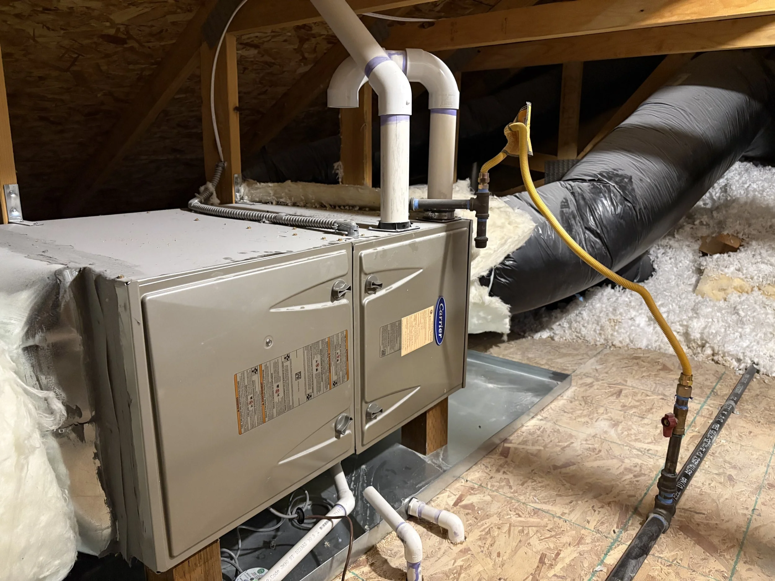 An HVAC furnace connected to air duct pipes and plumbing pipes in an attic with insulated walls and wooden framing.