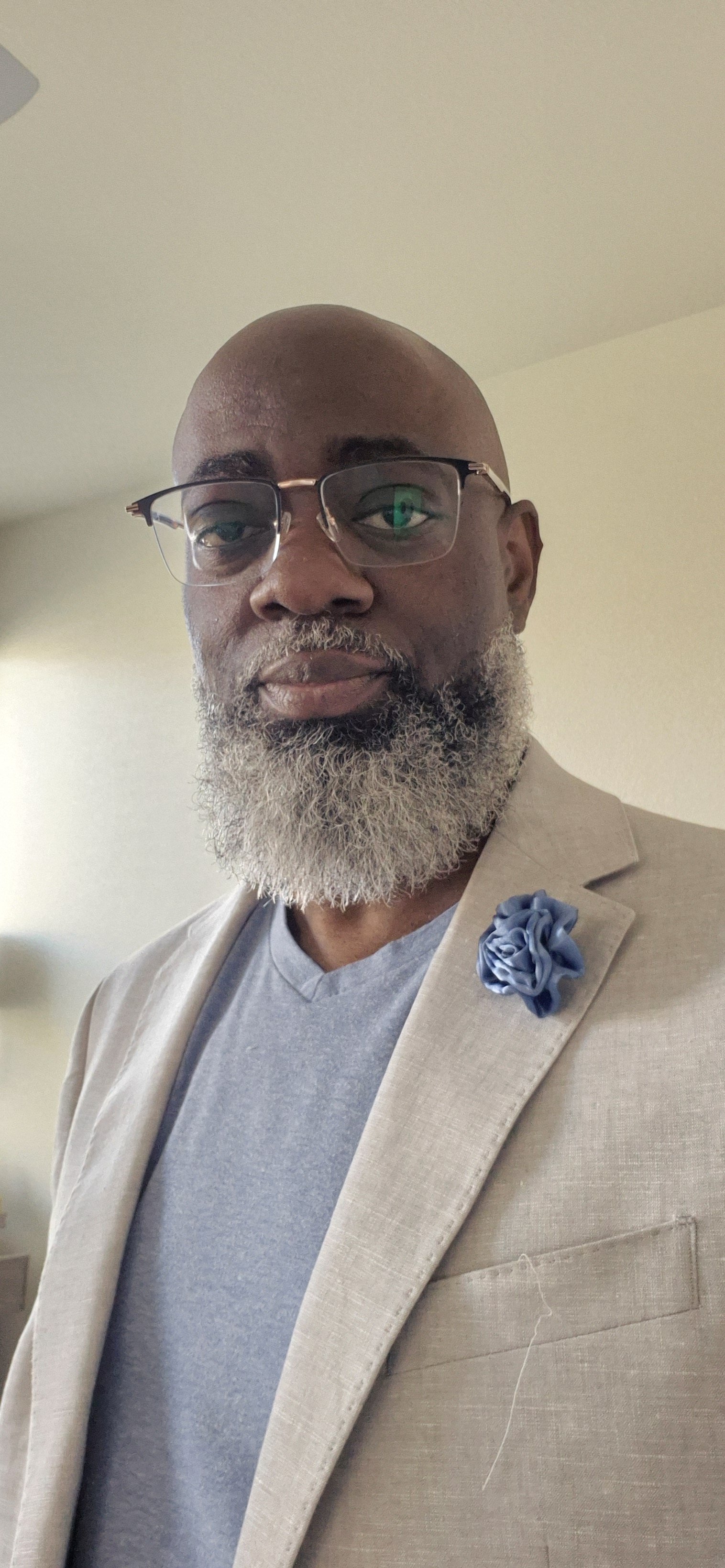 A man with glasses, a gray beard, and a bald head wearing a light beige blazer with a blue flower lapel pin, and a gray t-shirt.