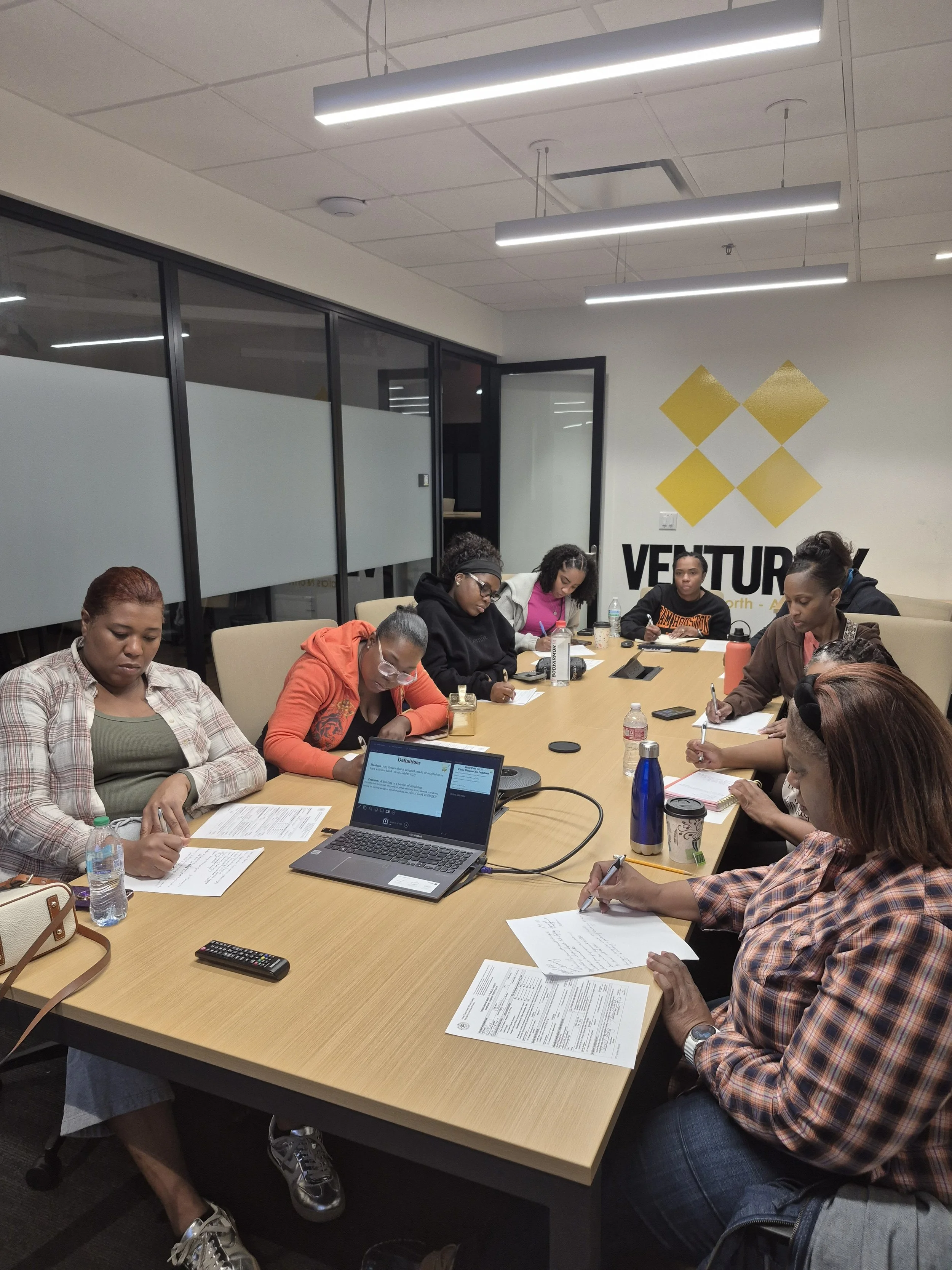A group of diverse women sitting around a conference table in a modern office, engaged in a meeting, with papers, notebooks, and drinks in front of them.