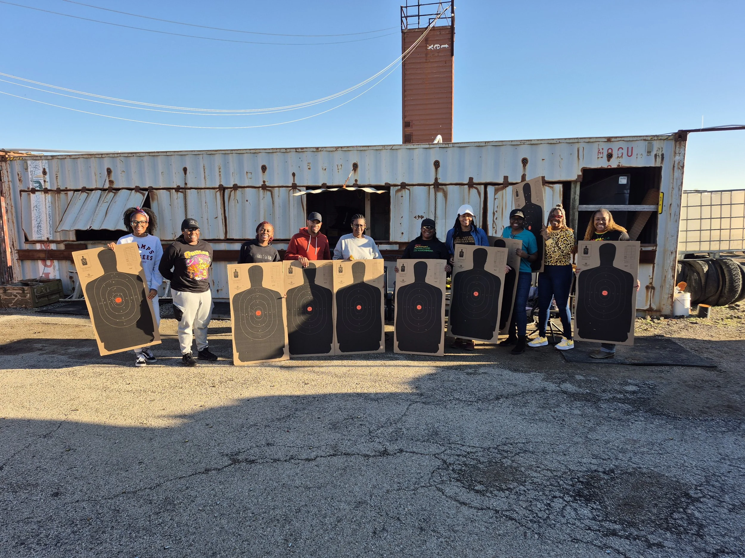 A group of nine diverse people standing outdoors in front of a rusted shipping container, holding large cardboard target boards with gun target markings. They are smiling, dressed casually, and appear to be participating in a shooting or training event on a sunny day.