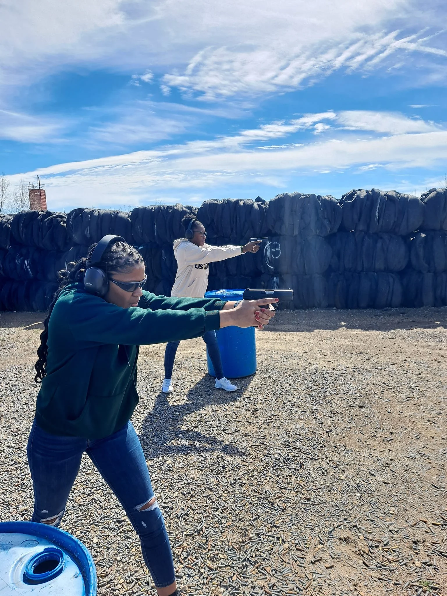 Three people at an outdoor shooting range aiming guns, with a large pile of shell casings on the ground and a blue sky with clouds overhead.