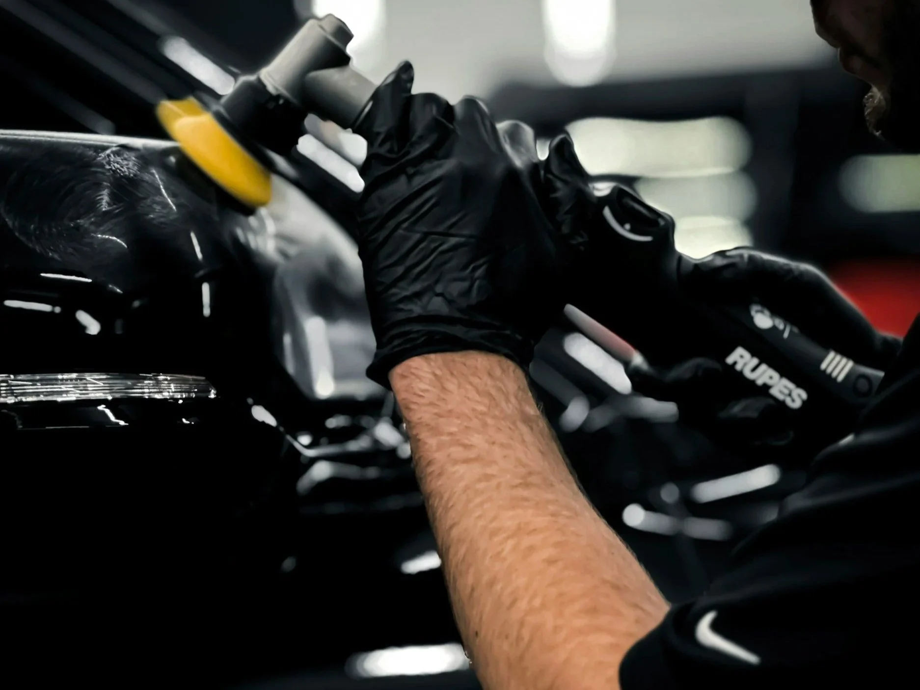 Close-up of a Person's hand wearing a black glove, using a yellow and black polishing tool on the body of a black car.