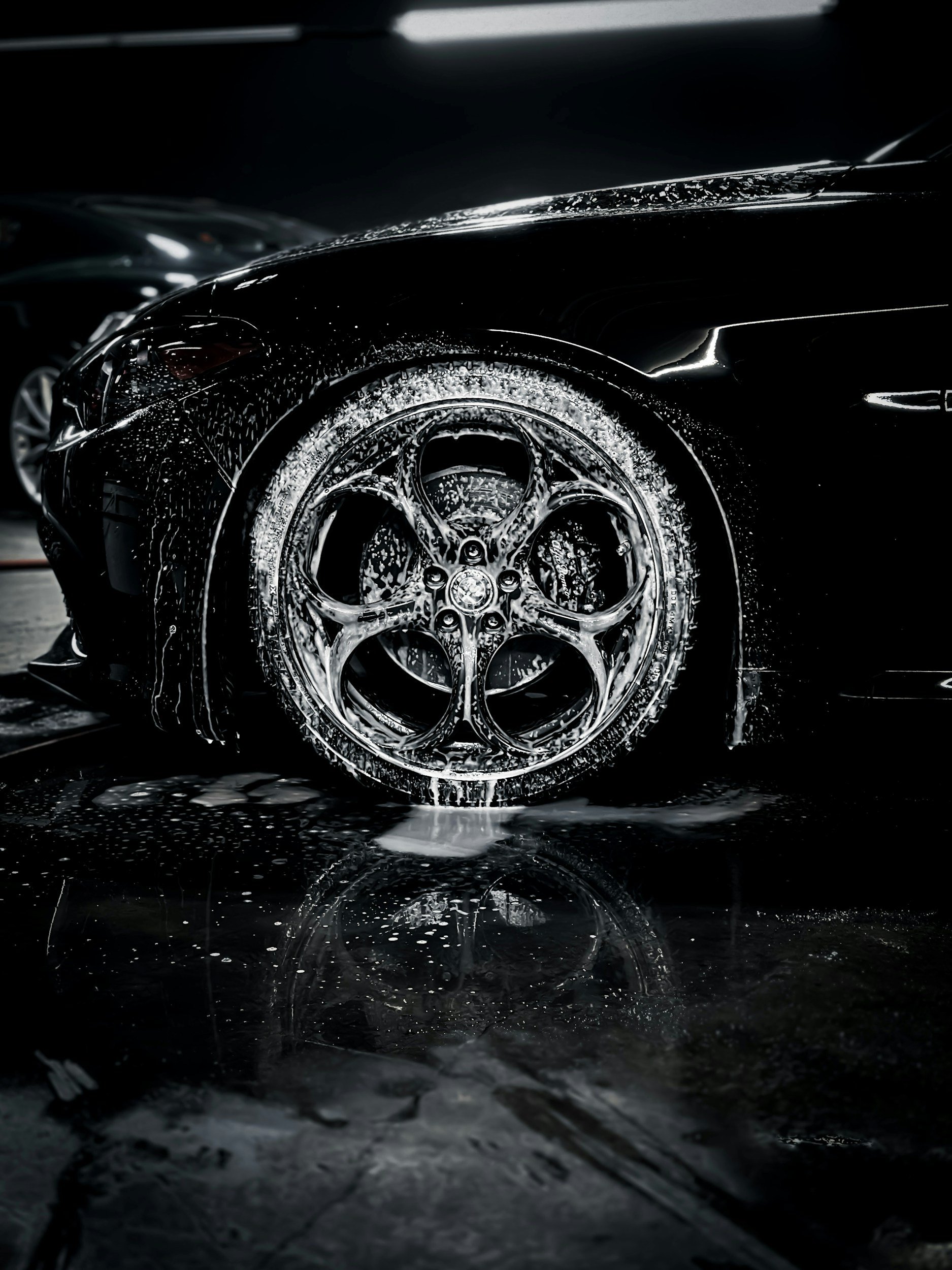 Close-up of a black sports car with water and soap suds on its body and wheel, in a dark setting.
