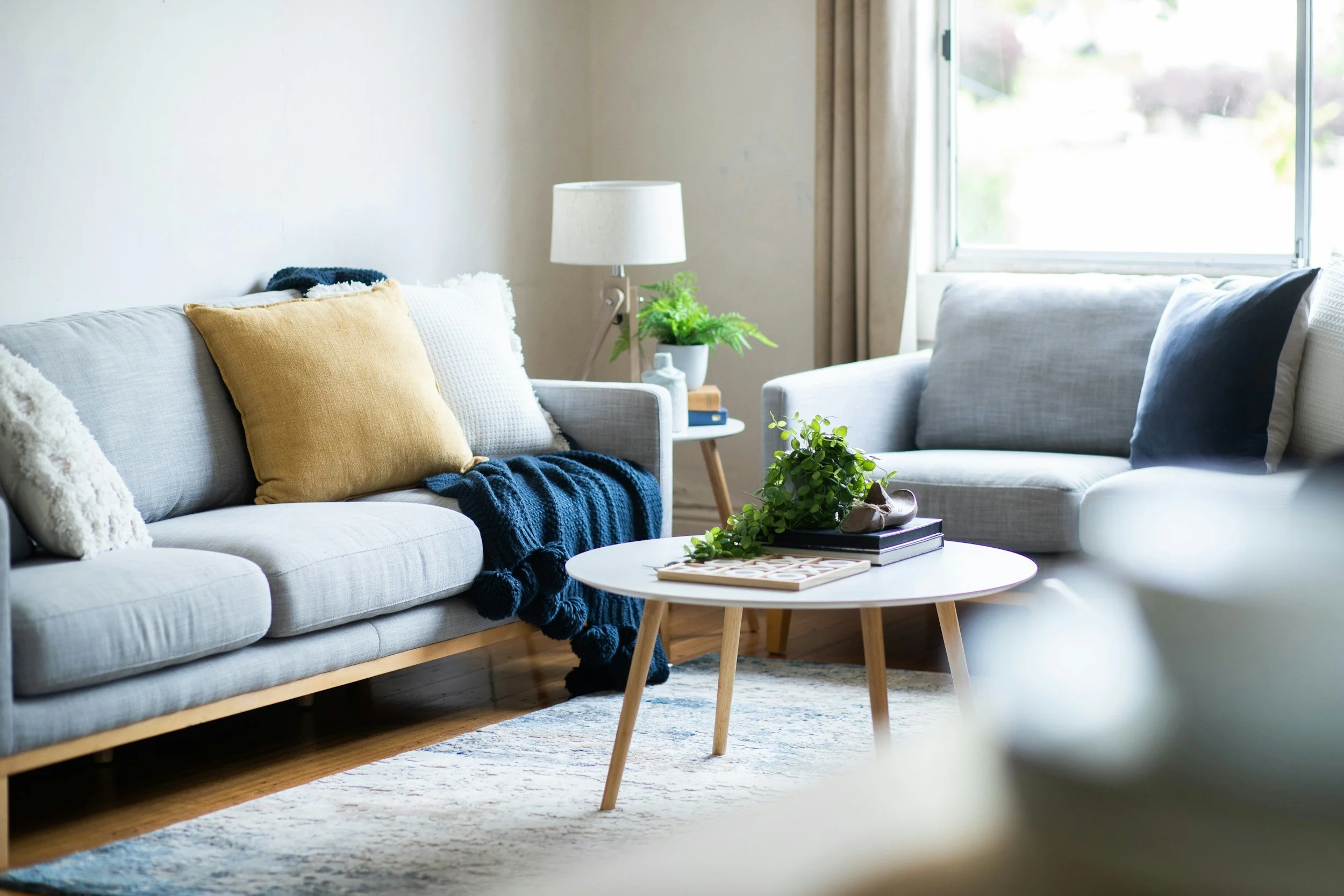 Bright living room featuring a light gray sofa with yellow, white, and dark gray pillows, a knit throw blanket, a round white coffee table with plants and books, a wooden side table with a white lamp and potted plant, and a large window with curtains letting in natural light.