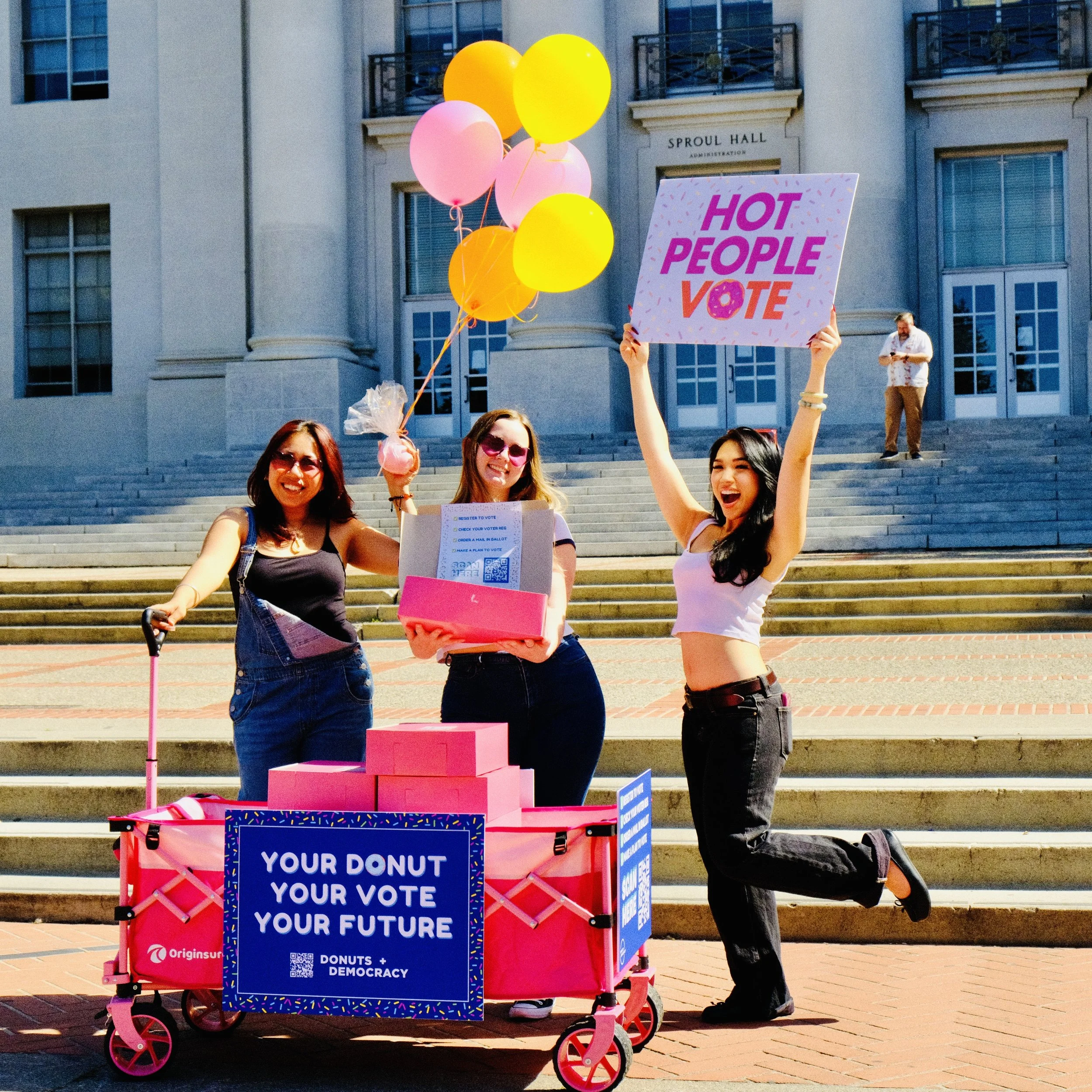 Three people with balloons, a donut wagon, and a "hot people vote" sign