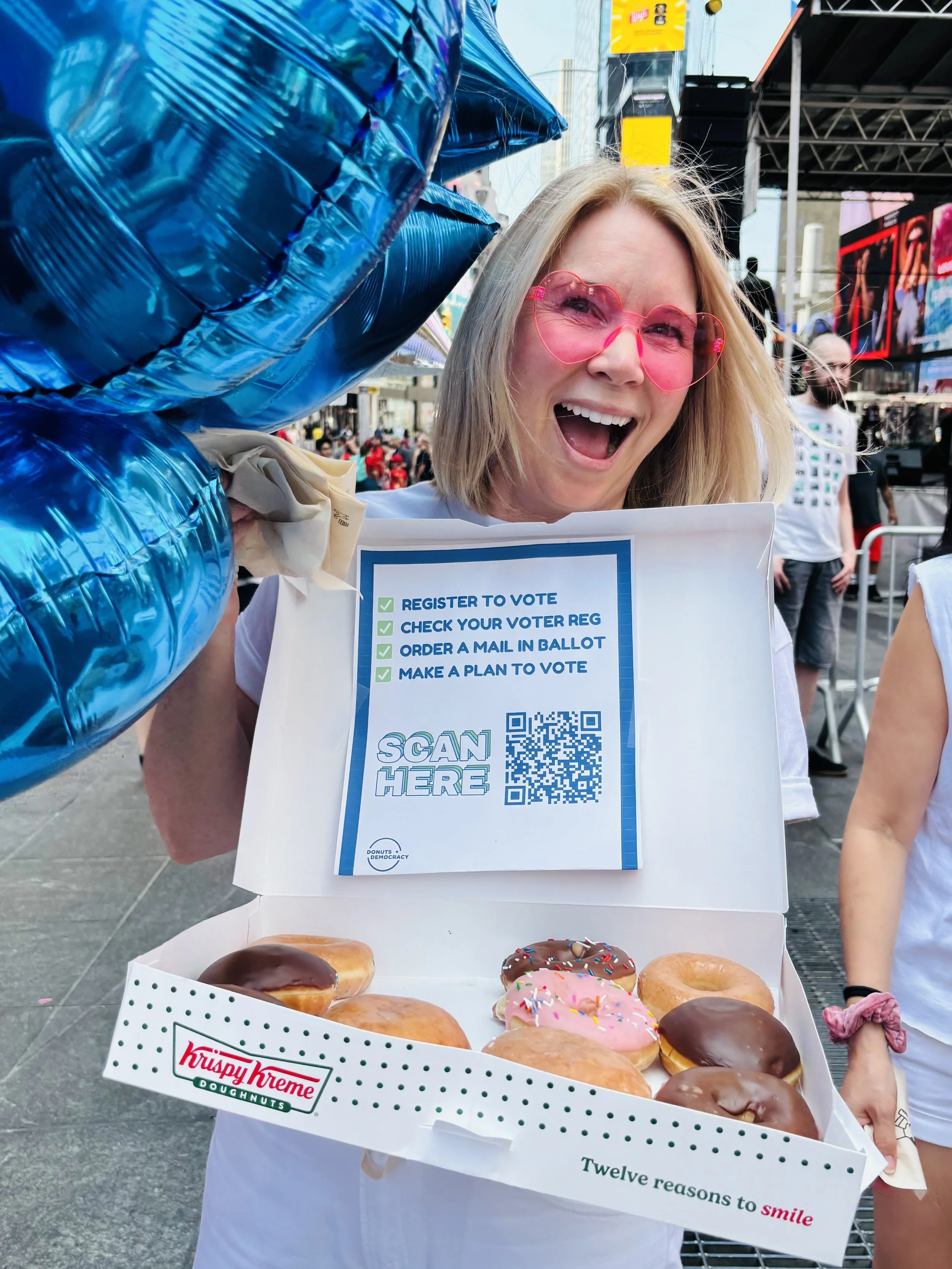 A person holding balloons, a donut box, and a sign to register to vote