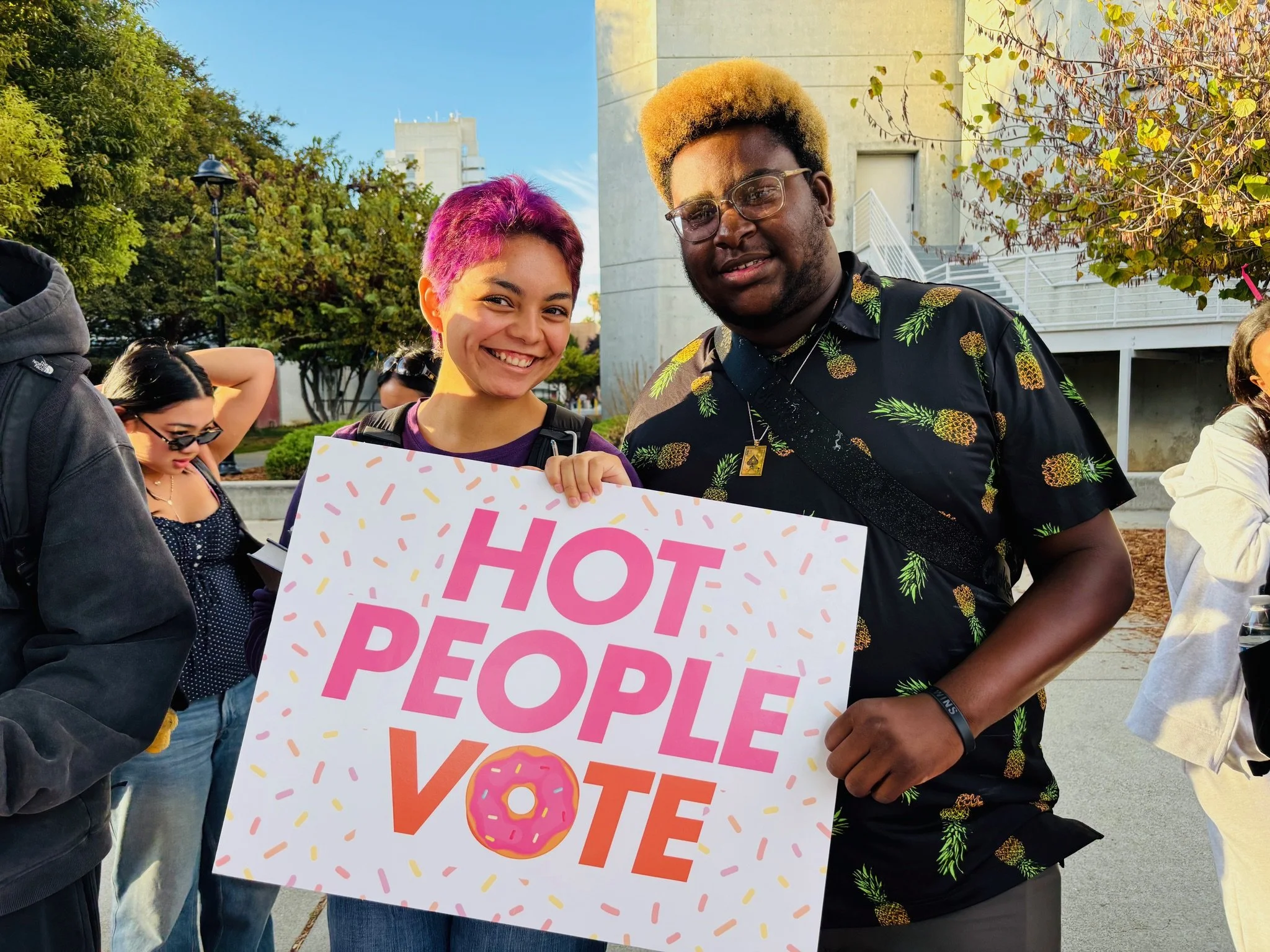 Two people with a sign that says "hot people vote"