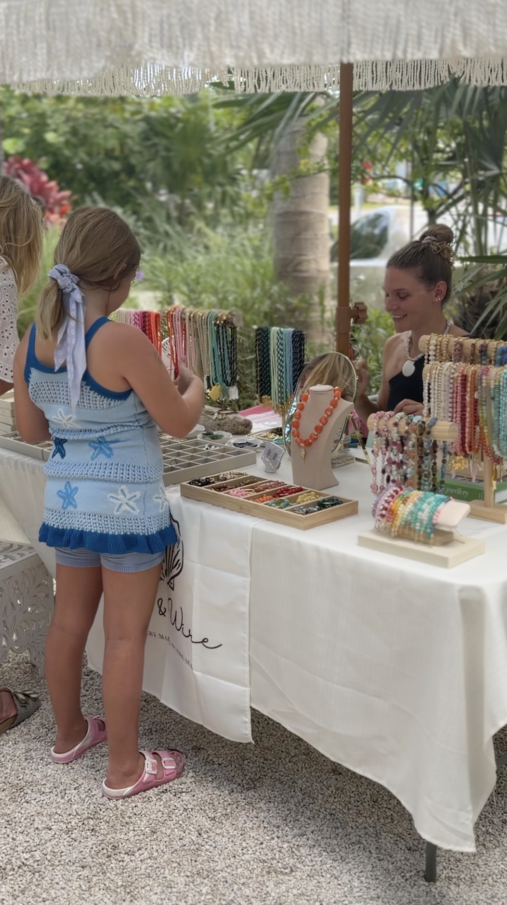 A woman selling jewelry at a market stall, and a girl looking at the jewelry, in an outdoor setting with greenery and palm trees in the background. The stall displays necklaces, bracelets, and other accessories.