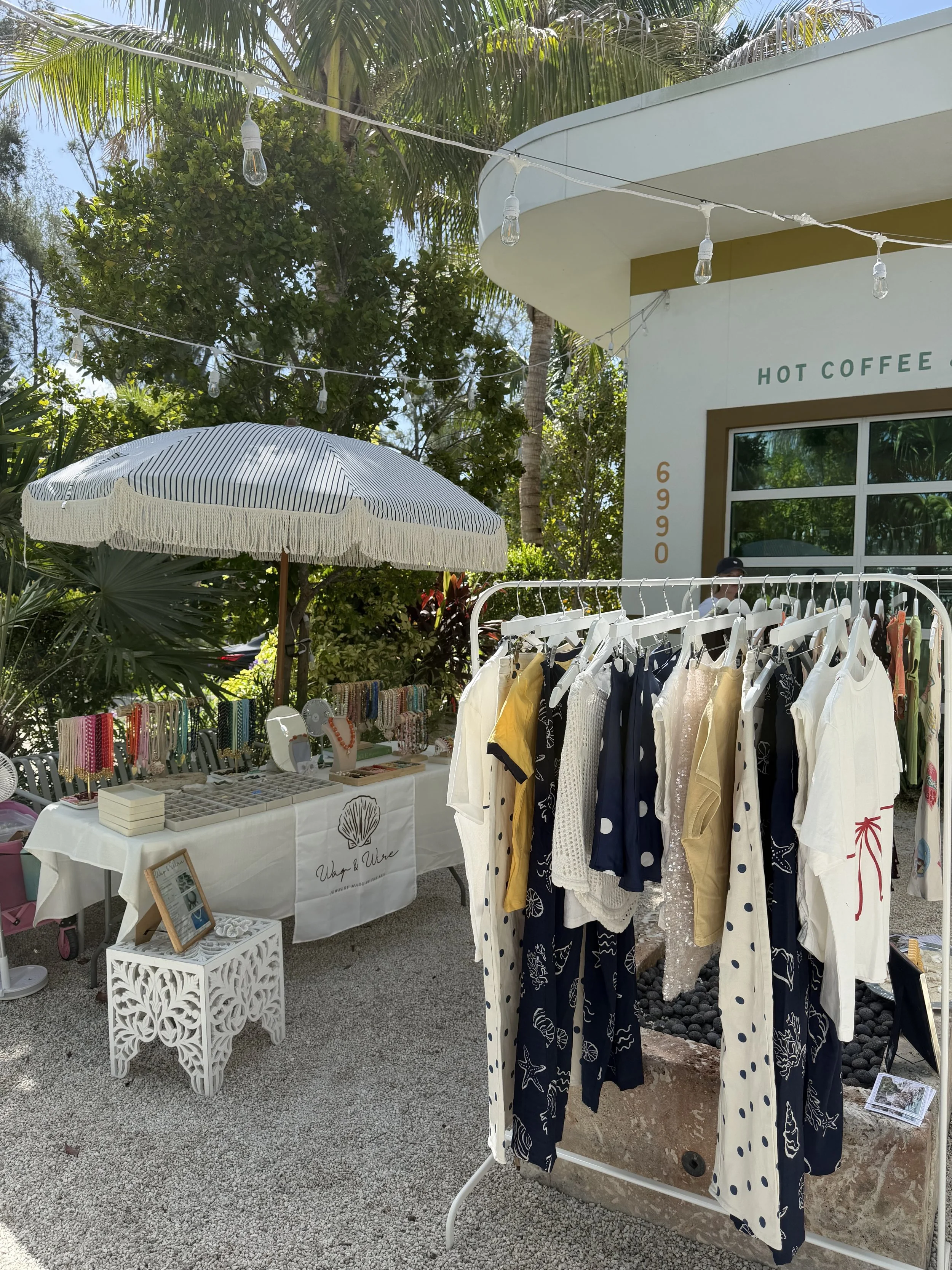 Outdoor market selling clothing and jewelry near a hot coffee stand with palm trees and string lights overhead.