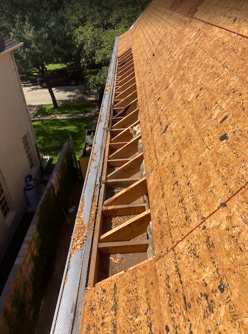 View of a roof under construction with exposed wooden rafters and plywood sheathing, overlooking a suburban street with trees and parked cars.