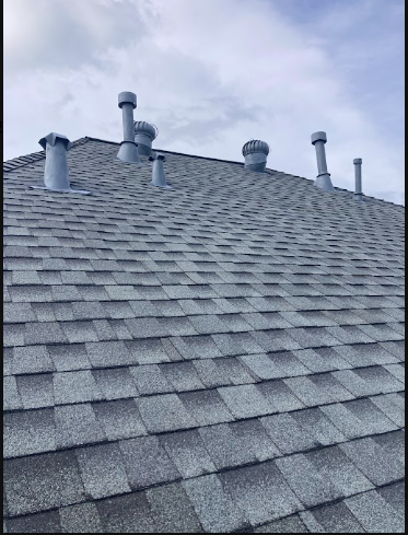 View of a gray shingled house roof with multiple vent pipes and a cloudy sky in the background.