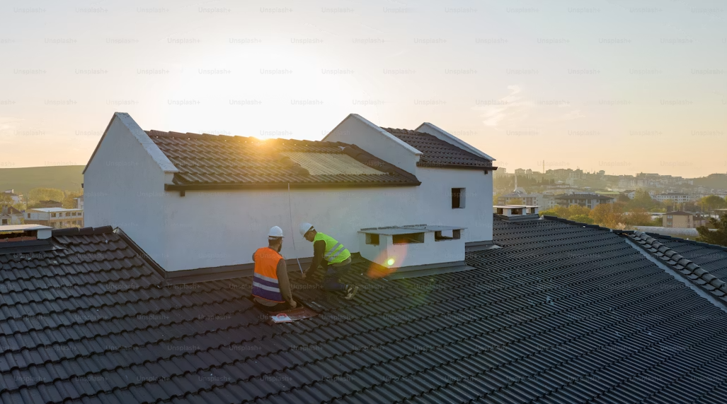 Two construction workers wearing safety gear and helmets working on a dark tiled roof at sunset.