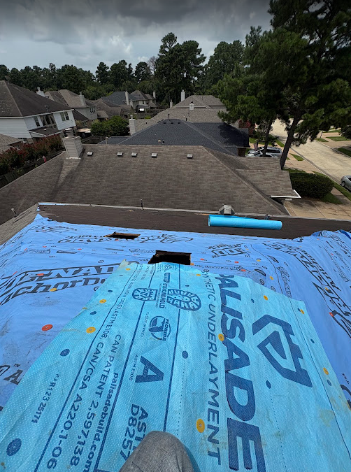 View from a roof under construction with blue insulation fabric, neighboring houses, trees, and a cloudy sky in the background.