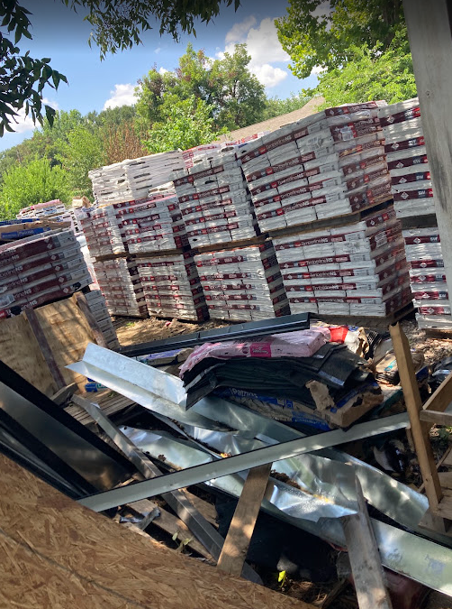 Stacks of construction materials, including bags of cement and metal sheets, outdoors on a sunny day with green trees in the background.