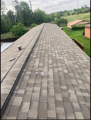 View of a shingled rooftop with a black cable running along the ridge, side vents, and a background of trees, grassy fields, and a neighboring house.