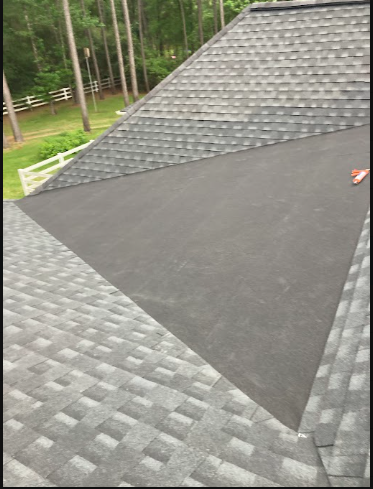 A roof with a section covered in new black asphalt shingles and another section in gray shingles with a textured pattern, surrounded by trees in the background.