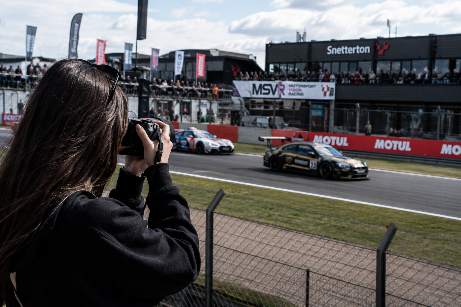 A woman with long brown hair, wearing a black hoodie and sunglasses on her head, taking a photo with a DSLR camera at a motorsport race track. Two race cars are on the track, while spectators watch from the grandstand and buildings labeled 'Snetterton' and 'MSVR' are in the background.