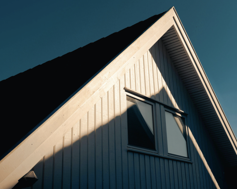 Close-up of a house with a triangular roof, light-colored siding, and a window, under a clear blue sky.