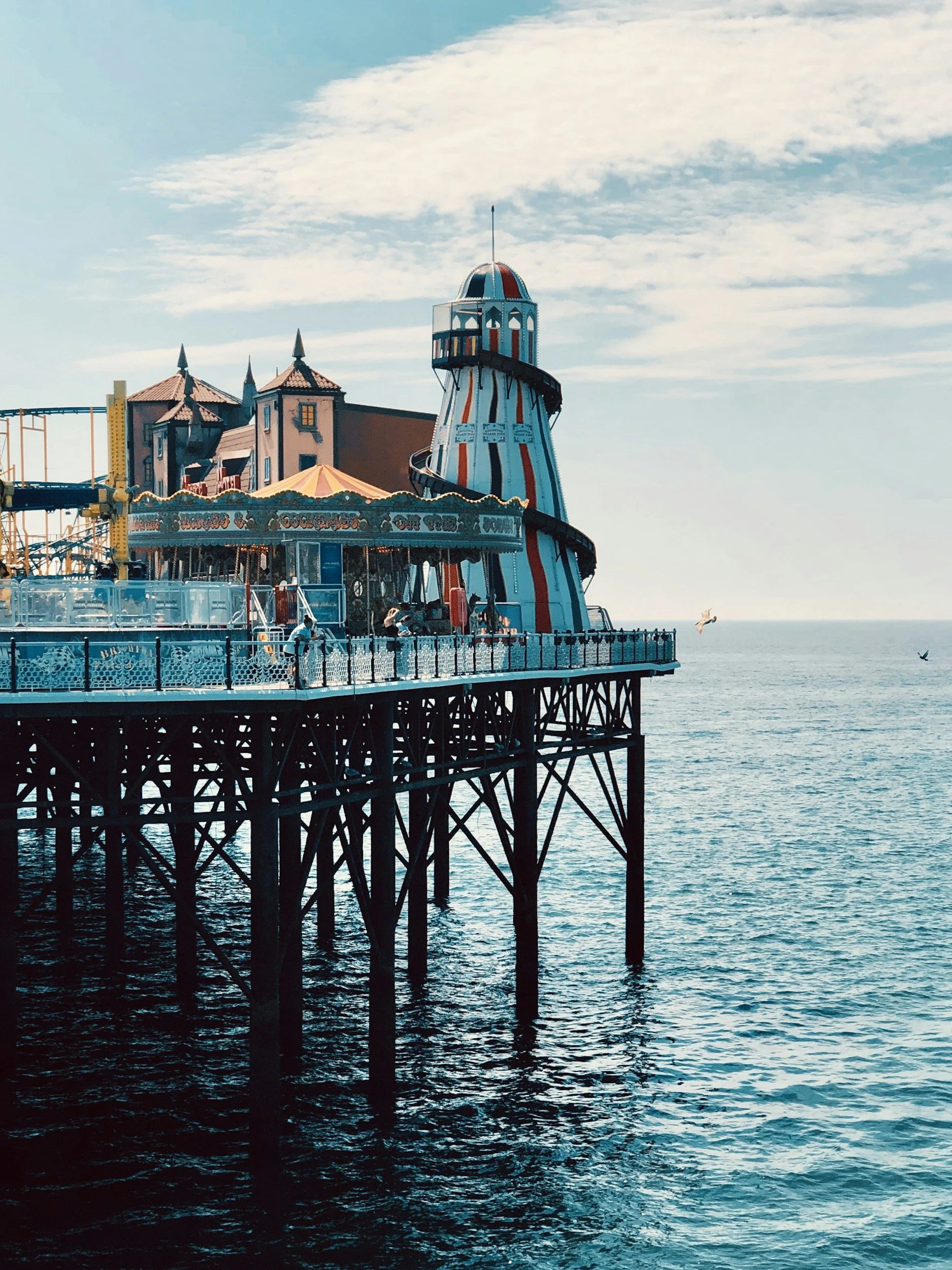 A seaside amusement park on a pier with a striped lighthouse, carousel, and a roller coaster visible, with seagulls flying overhead against a partly cloudy sky and calm ocean waters below.