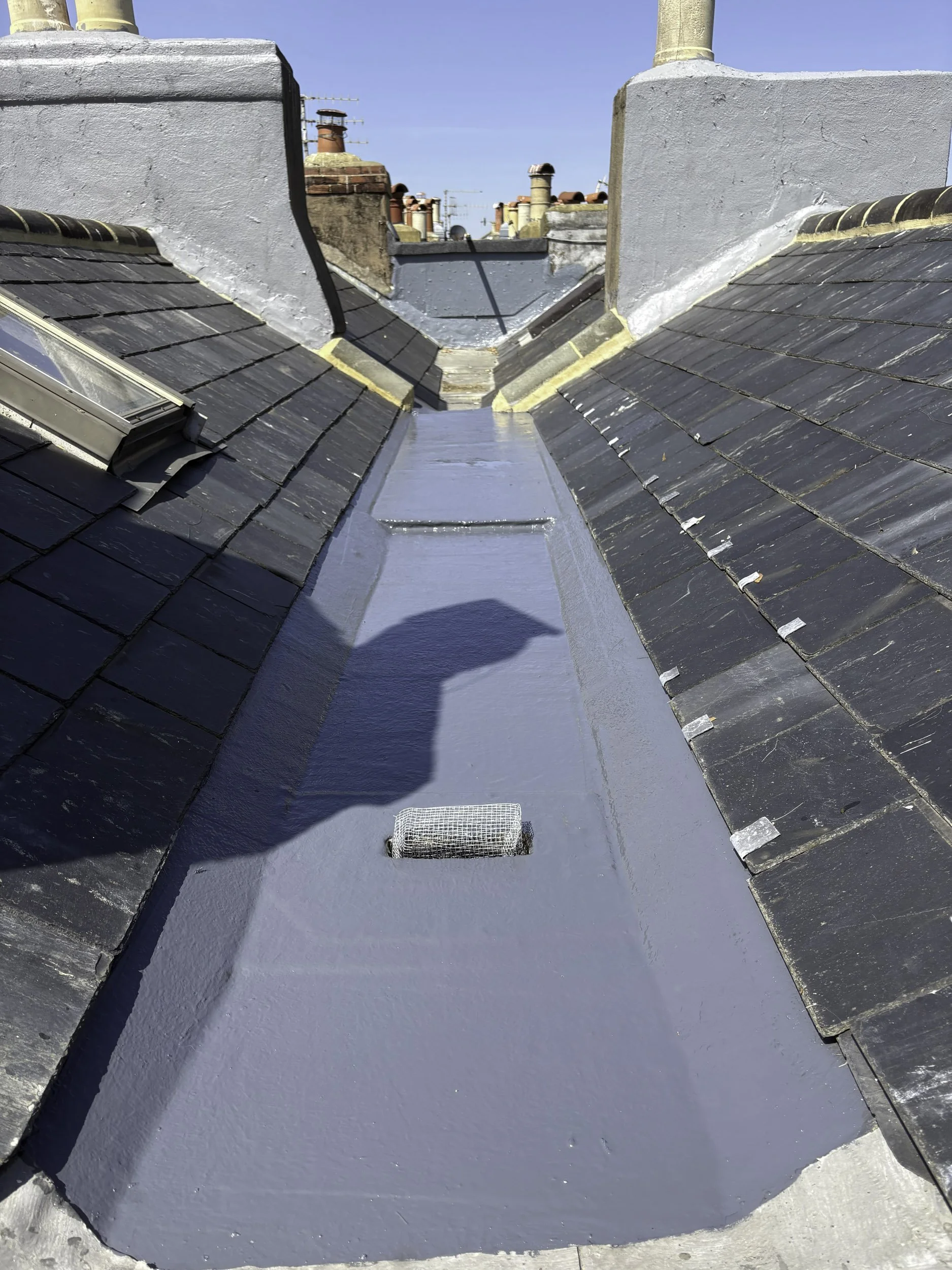 Flat roof with black shingles, white painted chimney walls, and a metal vent cover. In the background, multiple rooftop chimneys and antennas are visible under a clear blue sky.