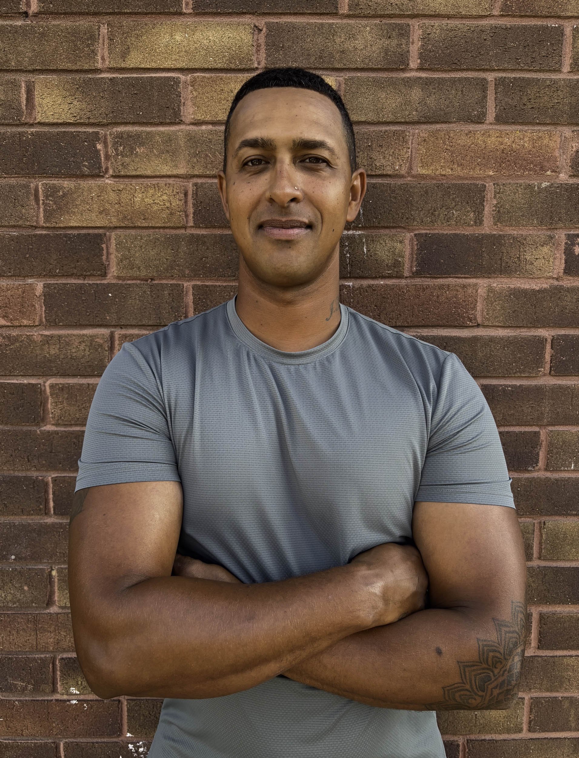 A young man with short dark hair and a nose piercing standing in front of a red brick wall, wearing a light blue athletic shirt, with arms crossed.