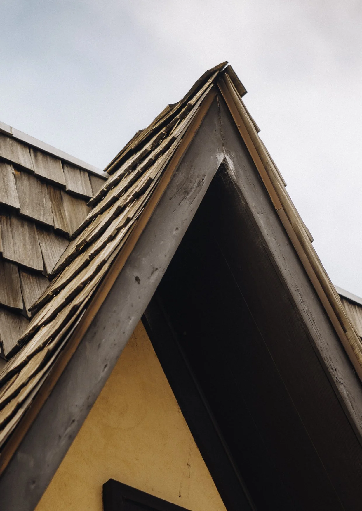Close-up of a roof gable with wooden shingles and a metal gutter, part of a house with an orange wall, under a cloudy sky.