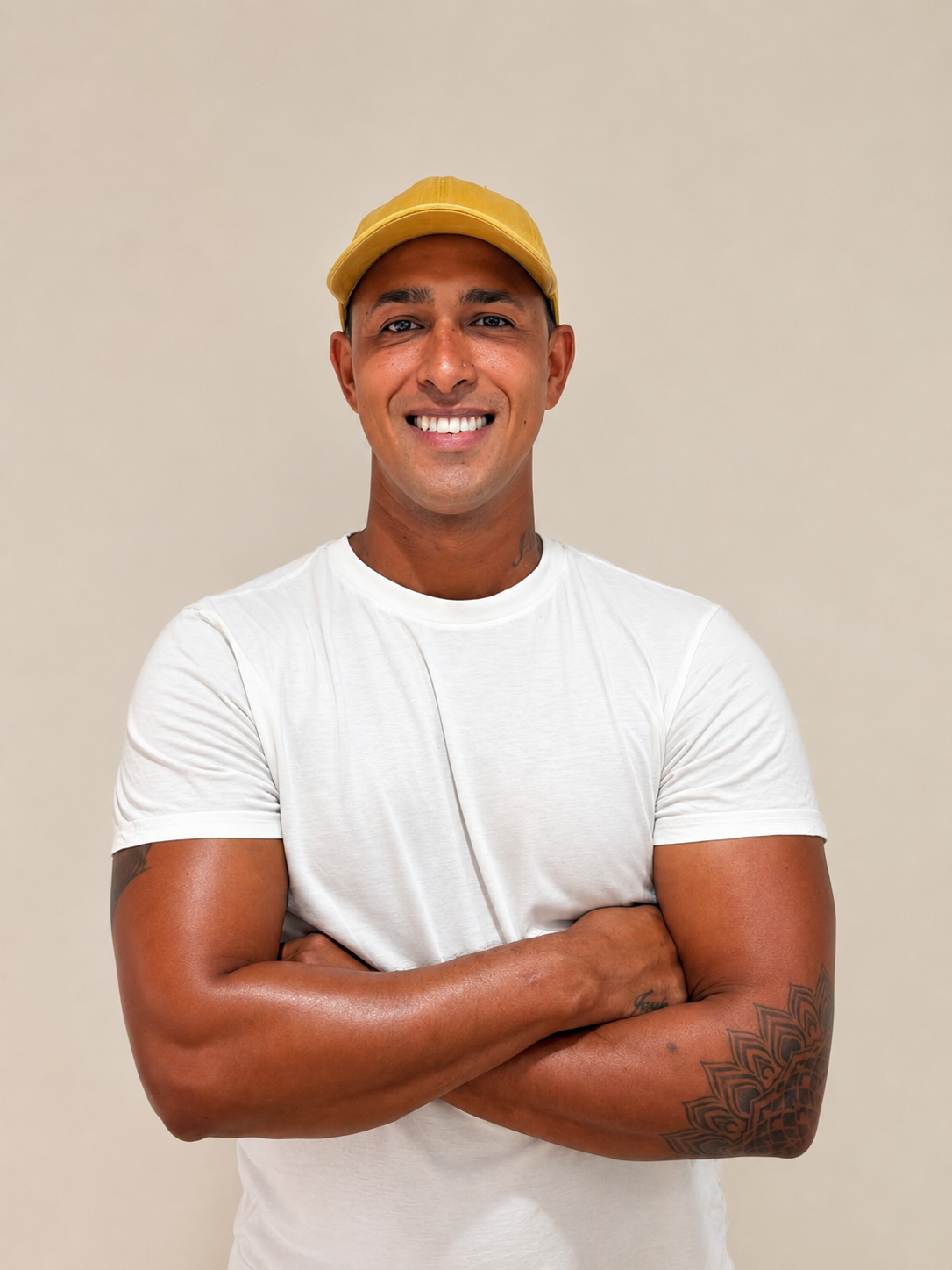 A smiling man in a yellow cap and white t-shirt, with crossed arms and tattoos on his left arm, standing against a plain beige background.