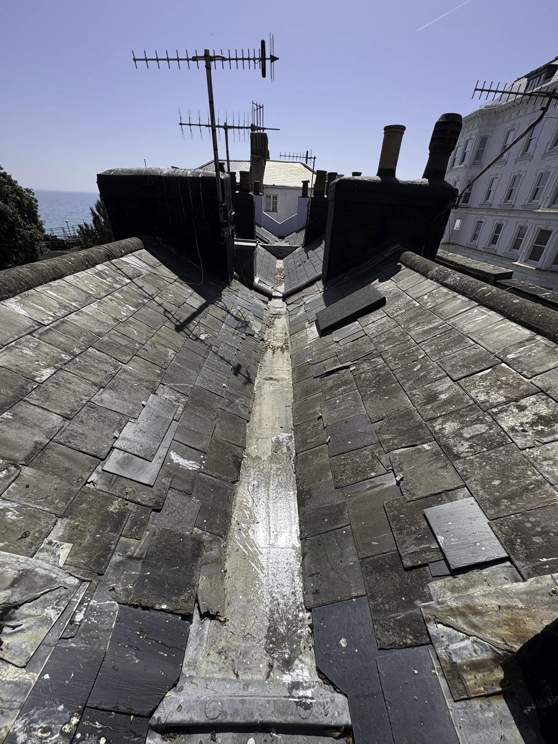 Close-up view of a rooftop with slate tiles, chimneys, rooftop antennas, and a clear blue sky with a faint contrail.