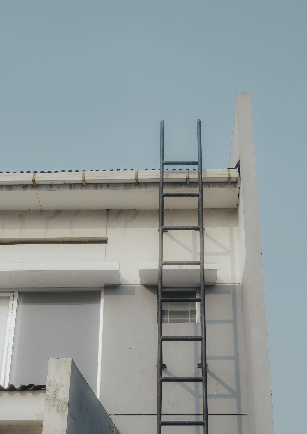 The image shows a white modern building with a black metal ladder leaning against the wall, reaching to the roof, under a clear blue sky.