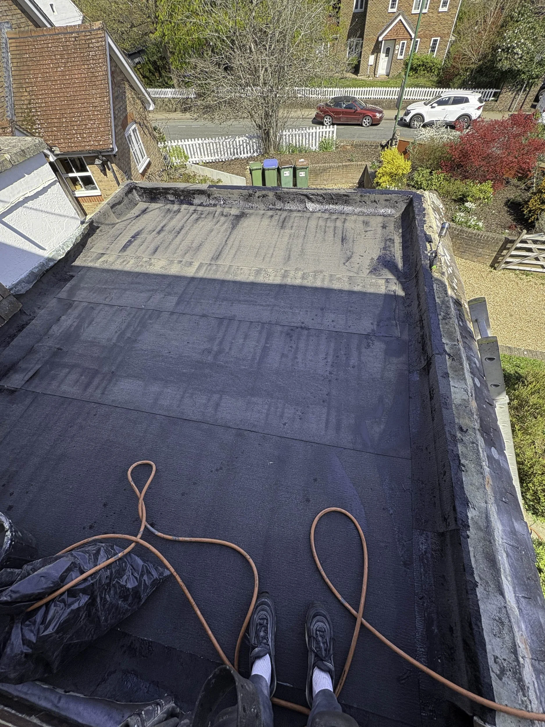 Person standing on a flat roof covered with black waterproofing material with orange extension cords. A plastic sheet bag is on the left. The view overlooks a suburban street with parked cars, trees, a white picket fence, and homes with brick walls and pitched roofs.