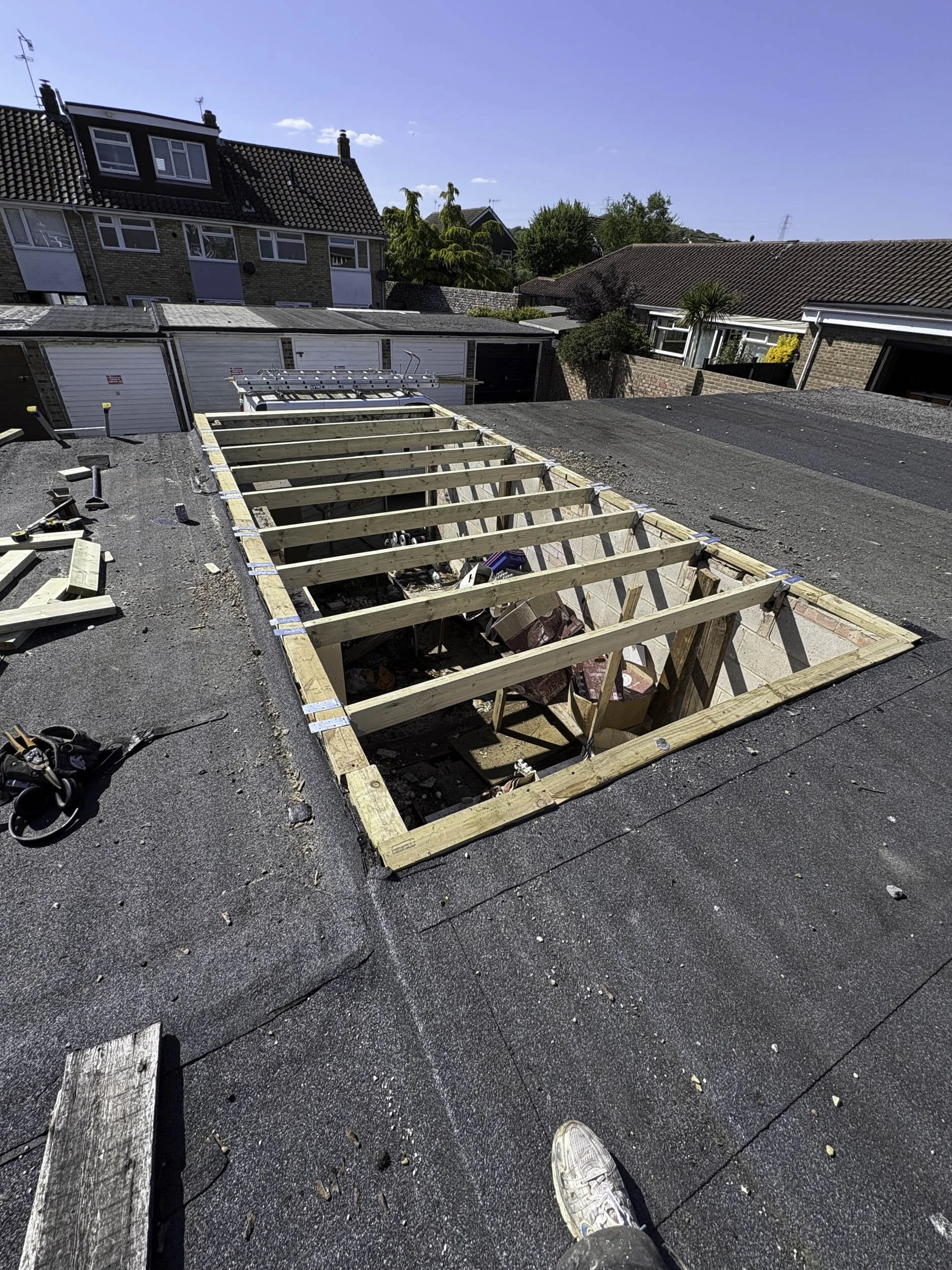 A rooftop under construction with wooden framing for a new section, construction tools and materials scattered around, neighboring houses in the background under a clear blue sky.