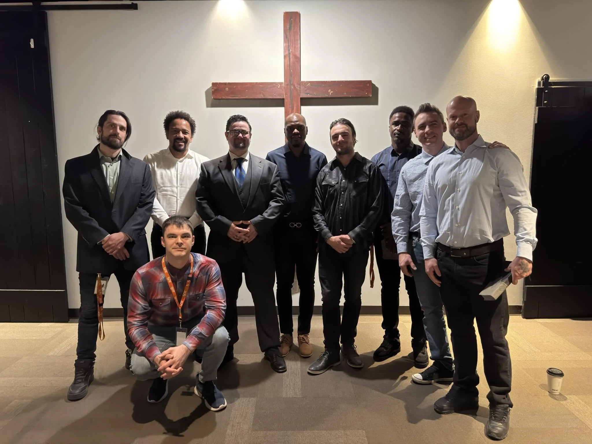 A group of nine men posed in front of a large wooden cross on a wall, some with their arms around each other, dressed in formal and casual attire, in an indoor setting.