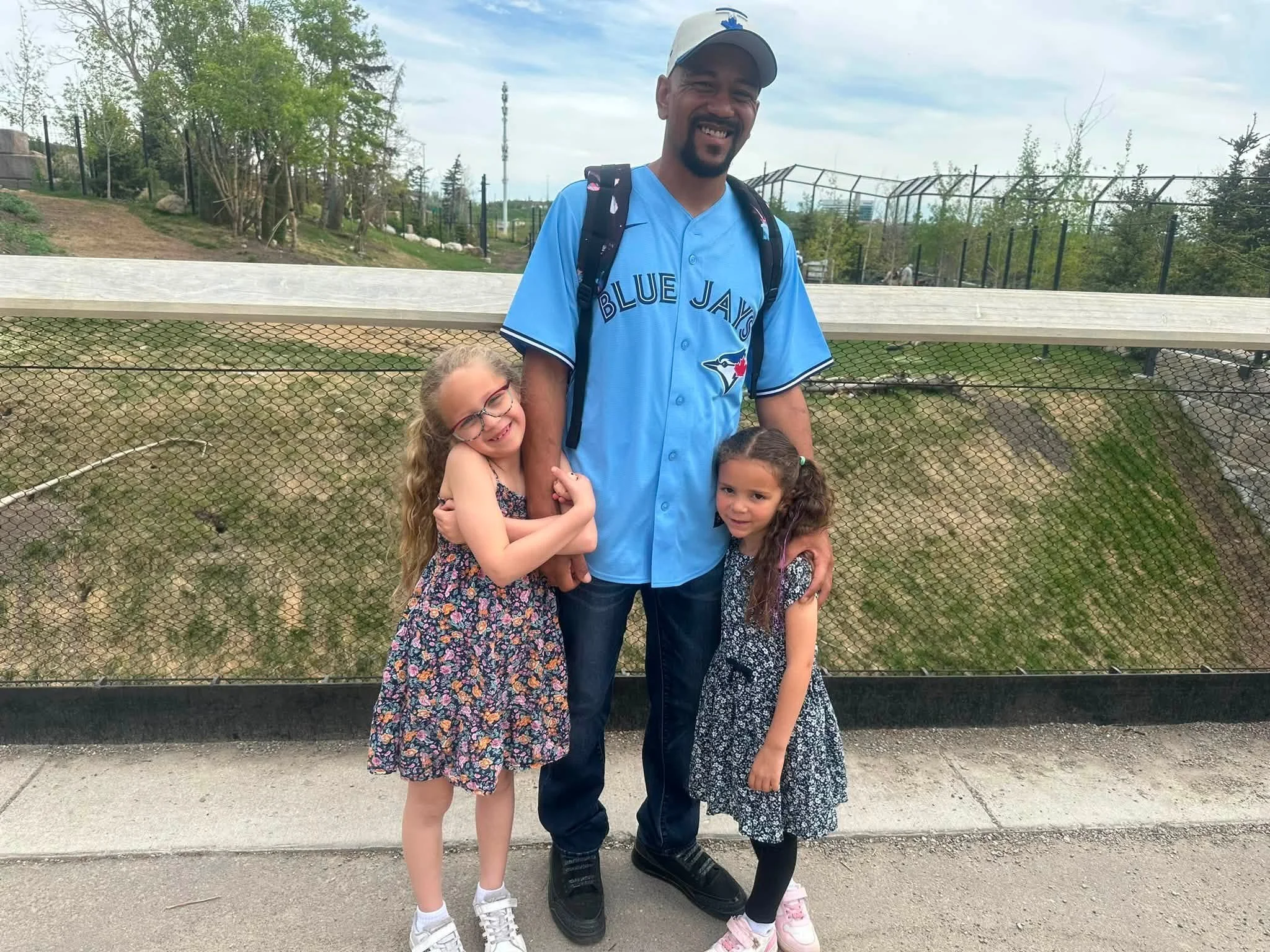 A man wearing a blue Toronto Blue Jays baseball jersey and a white cap standing on a sidewalk, smiling, with three young girls hugging him in front of a fence with trees and a park area in the background.