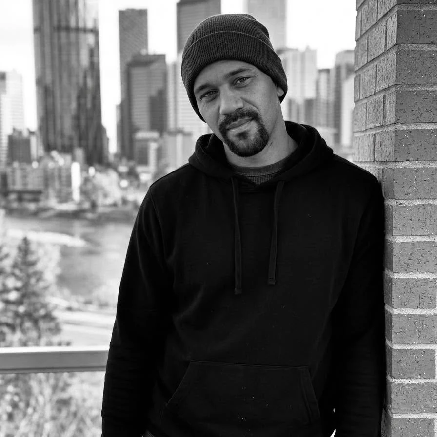 A man leaning against a brick wall outdoors with tall city buildings in the background, wearing a beanie and a black hoodie.