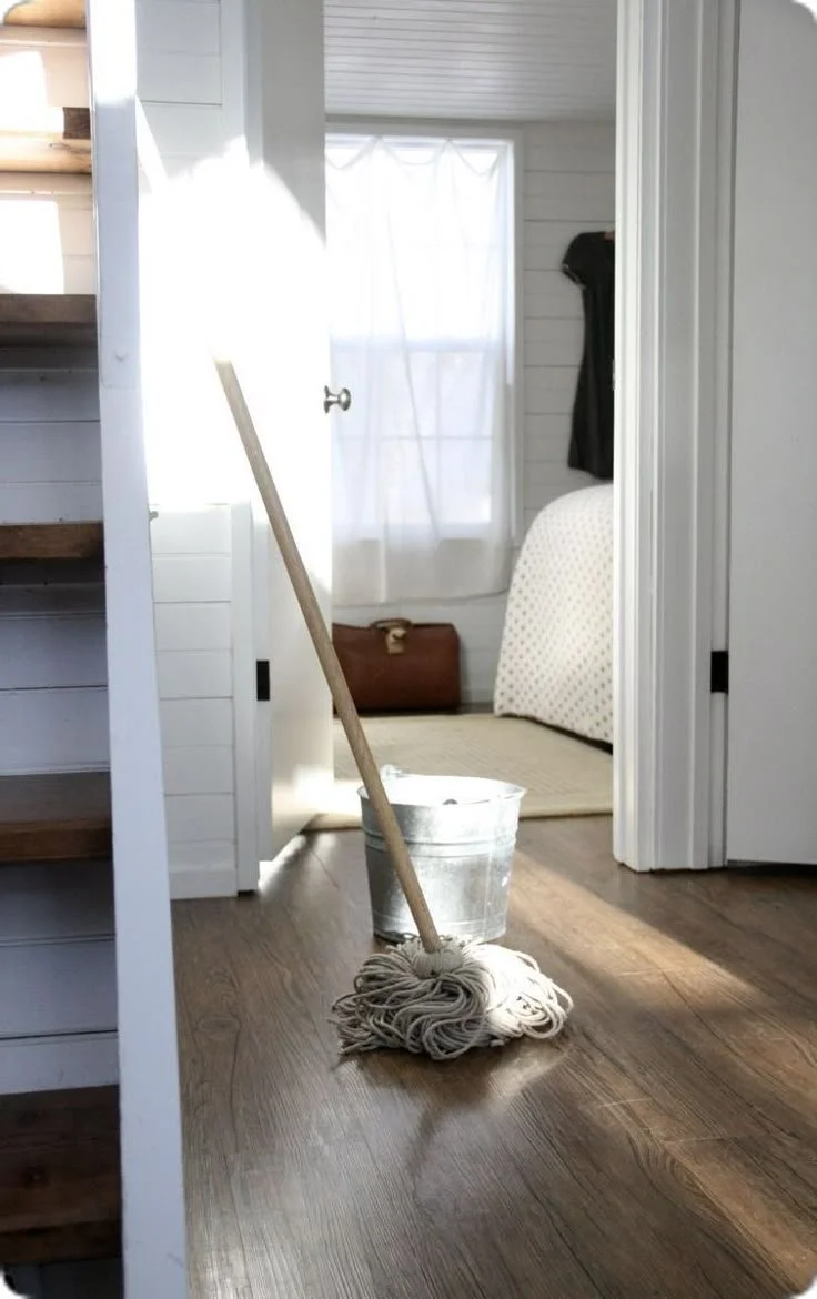A mop resting in a bucket on a wooden floor inside a house, with a doorway and a room with a window and a bed in the background.