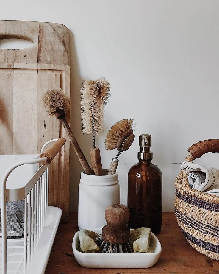 A collection of cleaning and household items on a wooden surface, including brushes in a white jar, a spray bottle, a woven basket with rolled towels, and a wooden cutting board in the background.